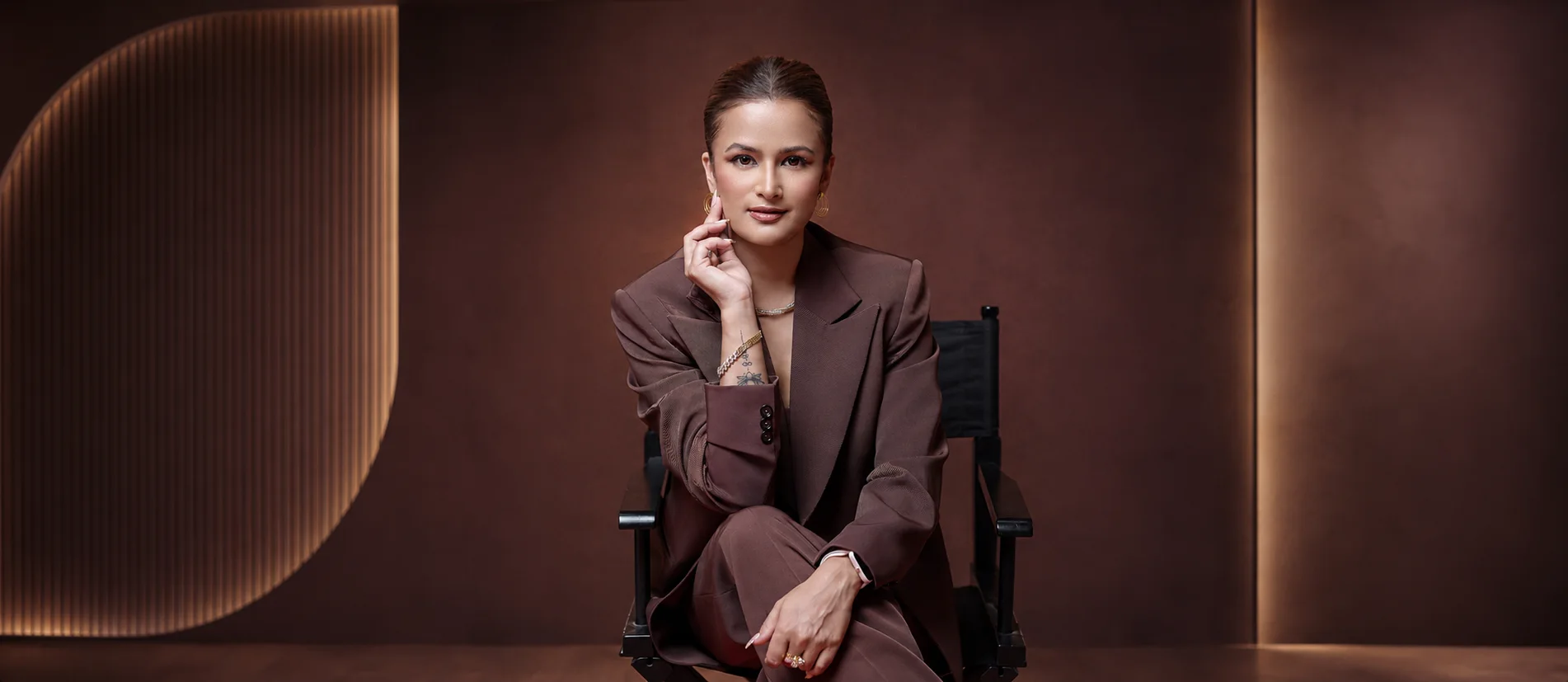Janu seated in a director's chair in a tailored brown suit against a warm studio backdrop